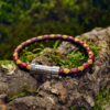 Ein geflochtenes Lederarmband mit roten und braunen Strängen und einer silbernen Schließe mit der Gravur AMPLI5 ruht auf einer moosbewachsenen Baumrinde im Freien.