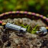 Ein geflochtenes Armband aus roten, gelben und schwarzen Strängen mit Metallverschlüssen, die an Klinkenstecker erinnern, ruht auf einer mit Moos bewachsenen Baumrinde.