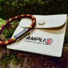 Ein geflochtenes rotes, schwarzes und gelbes Armband mit einem Metallverschluss ruht auf einem beigen Beutel mit der Aufschrift AMPLI5 Strength Balance Flexibility, der auf einer moosbewachsenen Rinde liegt.