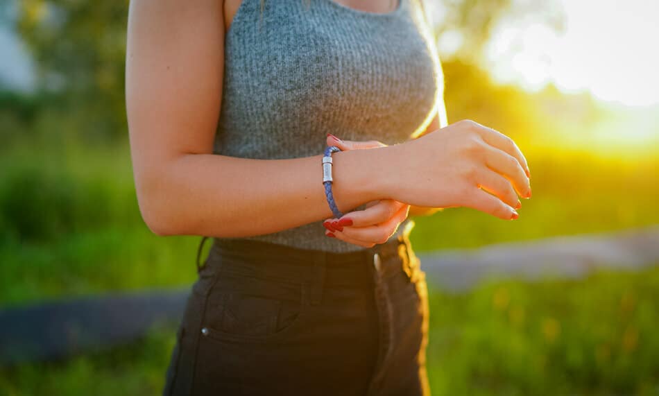 Eine Person, die ein graues ärmelloses Oberteil und eine schwarze Hose trägt, steht bei Sonnenuntergang im Freien und richtet ein blaues Armband an ihrem Handgelenk. Der Hintergrund ist grasbewachsen und unscharf, das Sonnenlicht erzeugt ein warmes Leuchten.