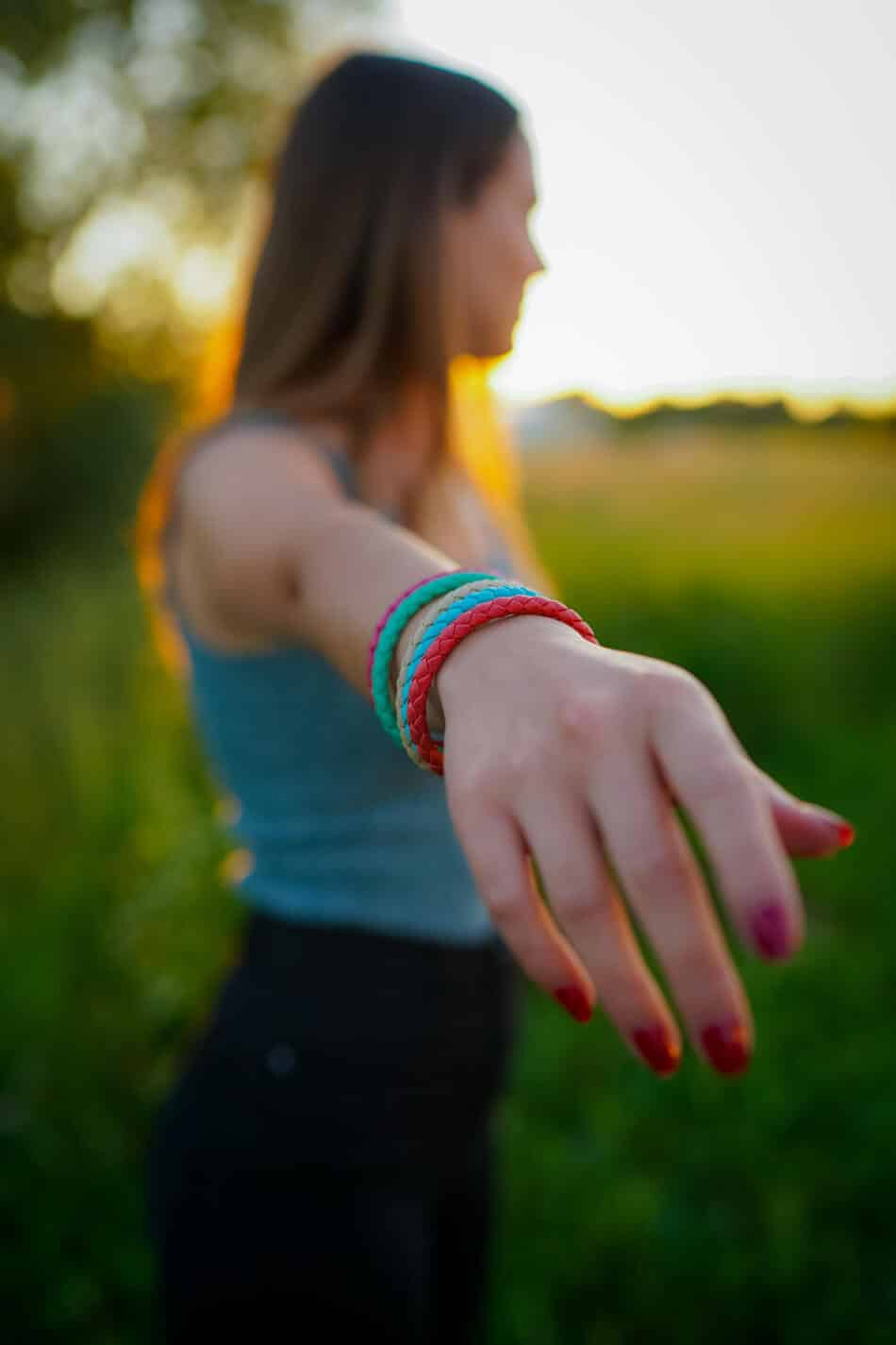Eine Frau mit langen Haaren steht bei Sonnenuntergang im Freien und streckt ihren Arm in Richtung der Kamera aus. Sie trägt bunte Armbänder und hat rot lackierte Nägel. Der Hintergrund besteht aus unscharfem Grün.