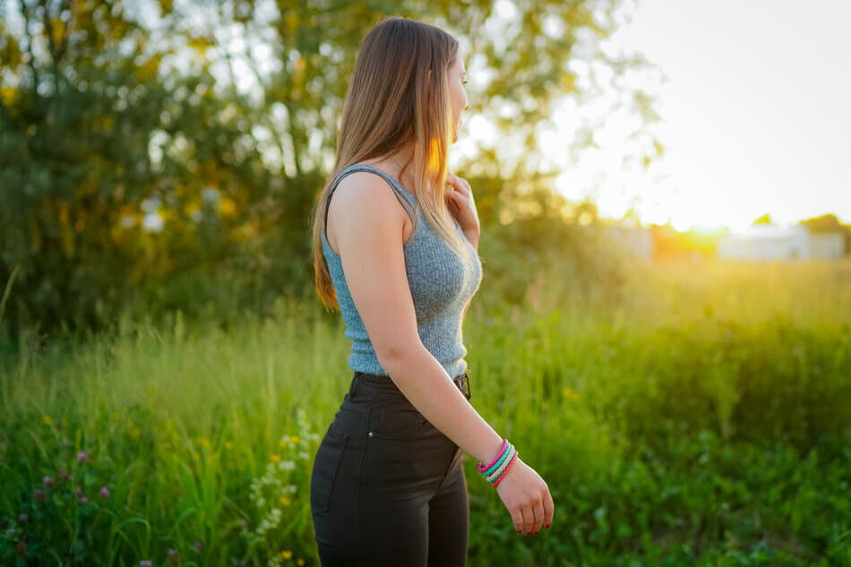 Eine Frau mit langen braunen Haaren, die ein graues Tanktop und eine schwarze Hose trägt, steht bei Sonnenuntergang im Freien auf einer Wiese und schaut zur Seite. Das Sonnenlicht fällt durch die Bäume im Hintergrund.