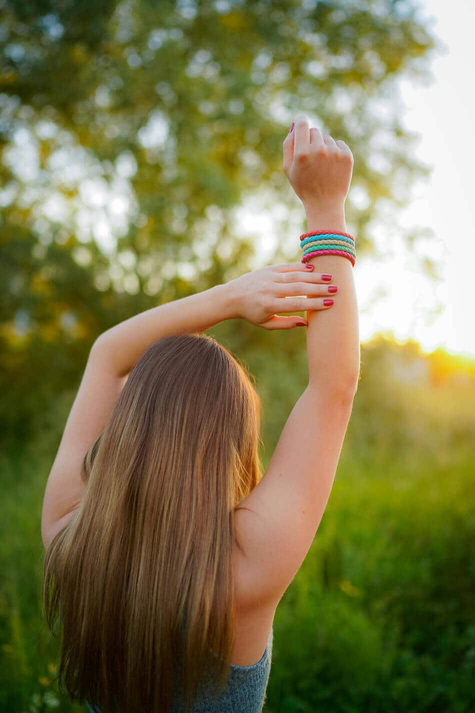Eine Frau mit langen braunen Haaren steht im Freien auf einer grünen Wiese und blickt von der Kamera weg. Sie hebt ihre Arme über den Kopf und trägt bunte Armbänder, während das Sonnenlicht durch die Bäume scheint.